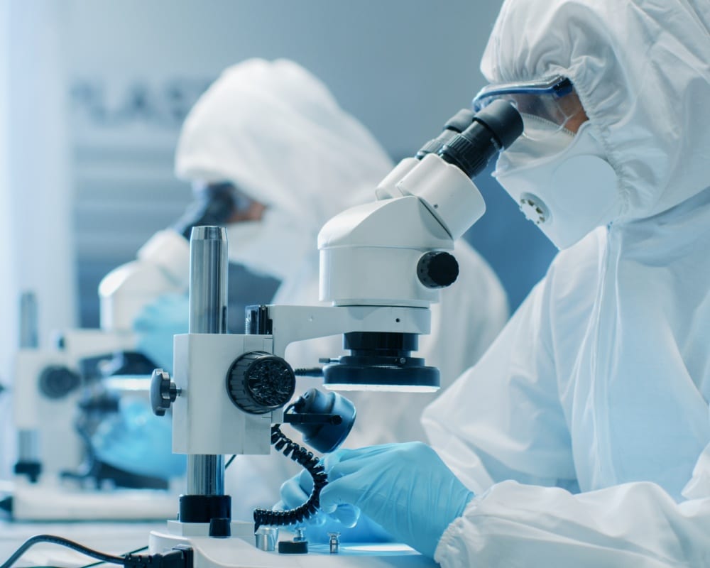 Two people wearing hazmat suits, safety glasses, and latex gloves, looking through microscopes