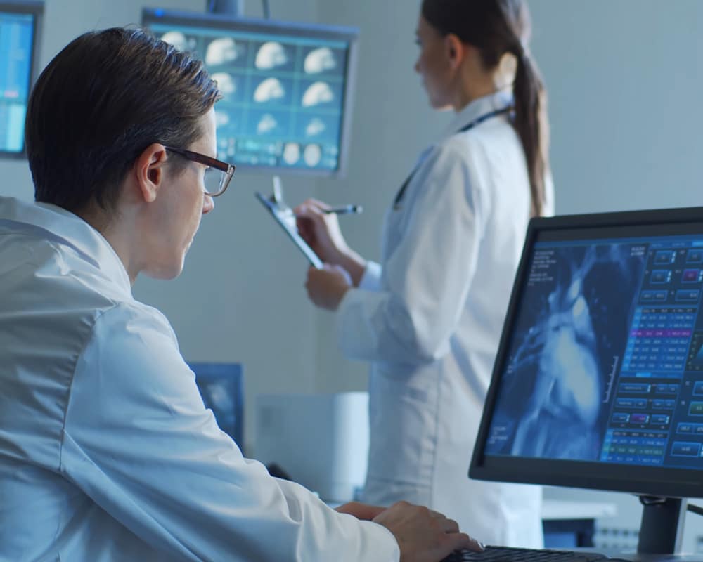 A medical professional in a lab coat analyses imaging data on a computer screen, while another professional in the background reviews patient records and scans in a clinical environment.