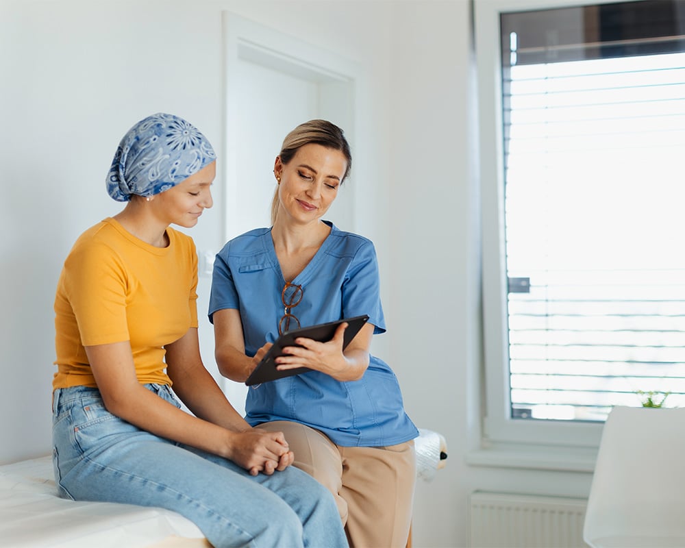 A healthcare professional in scrubs sits with a patient wearing a headscarf, holding a tablet and discussing information in a bright, supportive clinical setting.