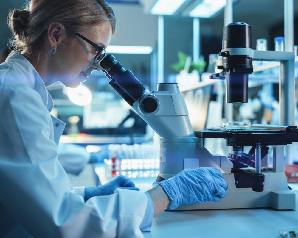 Woman scientist with blonde hair in pony tail wearing glasses looking through a microscope, wearing a lab coat and latex gloves